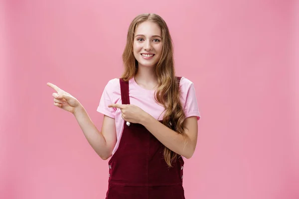 Beleefd en charismatisch mooi jong meisje met lang blond haar in corduroy trendy overall wijzend naar links alsof uitnodigen of tonen iets interessants lachend vriendelijk op camera over roze achtergrond — Stockfoto