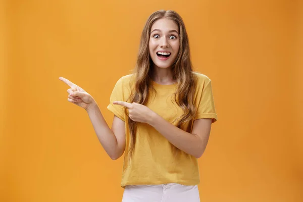 Retrato de la joven excitada maravillada y asombrada con cicatriz en el brazo y el pelo ondulado boca abierta en el entusiasmo y la alegría señalando izquierda divertido e interrogado ansioso por ver el espectáculo sobre la pared naranja — Foto de Stock