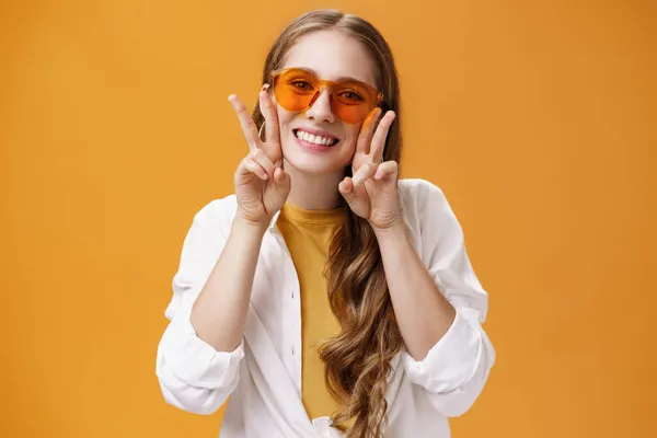 Elegante chica de fiesta entusiasta y carismática en gafas de sol de moda y blusa blanca sobre la camiseta que muestra gestos de paz cerca de la cara y sonriendo lindo en la cámara contra el fondo naranja —  Fotos de Stock