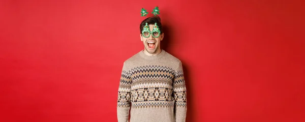 Πορτρέτο του έκπληκτος και ενθουσιασμένος όμορφος άντρας σε γυαλιά κόμμα και το χειμώνα πουλόβερ, γιορτάζει τα Χριστούγεννα και να διασκεδάζουν, στέκεται ενάντια στο κόκκινο φόντο — Φωτογραφία Αρχείου