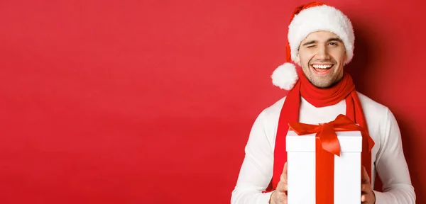 Έννοια των χειμερινών διακοπών, τα Χριστούγεννα και τον τρόπο ζωής. Όμορφος αναιδής άντρας με καπέλο και μαντήλι, που κρατάει το παρόν και χαμογελάει, κλείνει το μάτι στην κάμερα, στέκεται πάνω από το κόκκινο φόντο — Φωτογραφία Αρχείου