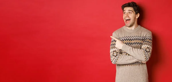 Portrait of attractive caucasian man celebrating new year, winter holidays, pointing finger and looking left with amazed expression, showing christmas advertisement, standing over red background — Stock Photo, Image