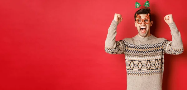 Porträt eines begeisterten und glücklichen Mannes mit Partybrille, Winterpullover, Freudenschreien und erhobenen Händen, Gewinn an Weihnachten, Erreichen des Neujahrsziels, stehender roter Hintergrund — Stockfoto