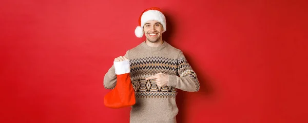 Conceito de férias de inverno, ano novo e celebração. Bonito homem sorridente preparou presentes para as crianças, apontando para bolsa de Natal, de pé sobre fundo vermelho — Fotografia de Stock