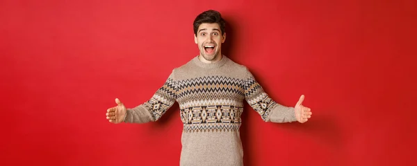 Retrato de homem feliz atraente em camisola de Natal mostrando grande presente, mãos estendidas lateralmente como se segurando grande caixa, de pé sobre fundo vermelho — Fotografia de Stock