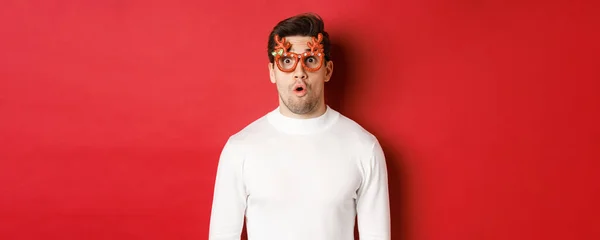 Close-up de homem surpreso e impressionado em suéter branco, óculos de festa, dizendo uau e olhando espantado para a câmera, de pé sobre fundo vermelho — Fotografia de Stock