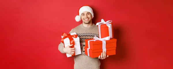 Концепція зимових свят, нового року та святкування. Портрет милого усміхненого чоловіка приготував подарунки на Різдво, тримаючи подарунки і дивлячись на сердечко камери, червоний фон — стокове фото