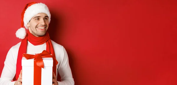 겨울 휴가, 크리스마스, 생활 양식의 개념. 산타 모자와 스카프를 착용하고 새해 선물을 들고 오른쪽을 바라보며 웃고 빨간 배경 위에 서 있는 매력적 인 남성의 근접 사진 — 스톡 사진
