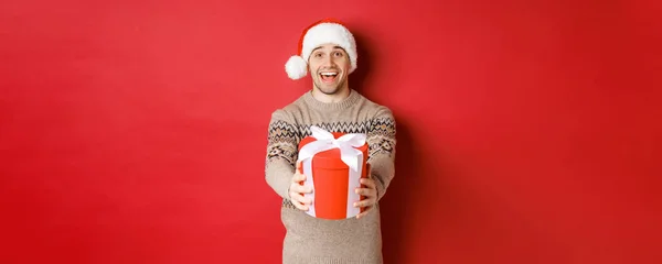 Imagem do homem feliz dizendo Feliz Natal e dando-lhe um presente na caixa, sorrindo alegre, comemorando férias de inverno em chapéu de santa e camisola xmas, de pé sobre fundo vermelho — Fotografia de Stock
