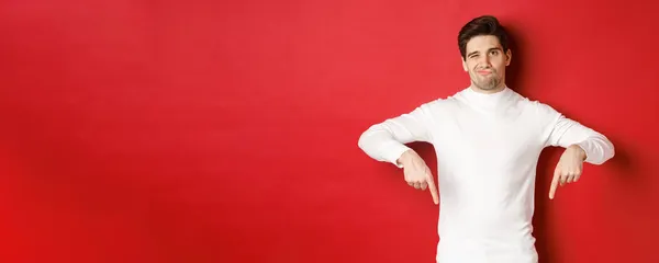 Retrato de atraente, jovem cético em suéter branco, risonho e apontando os dedos para baixo, discordar e não gostar de algo, de pé sobre fundo vermelho — Fotografia de Stock