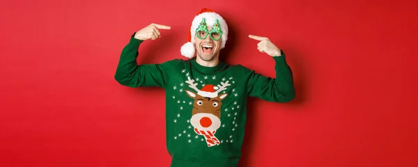 Image d'un gars souriant excité pointant vers les lunettes de fête et célébrant le nouvel an, se tenant joyeux dans un pull vert et un chapeau de Père Noël sur fond rouge — Photo