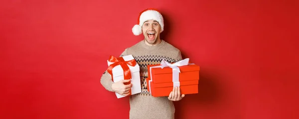 Konzept der Winterferien, Neujahr und Feier. Bild eines erstaunten und glücklichen Mannes in Pullover und Weihnachtsmütze, der Weihnachtsgeschenke in der Hand hält und vor Freude schreit, roter Hintergrund — Stockfoto