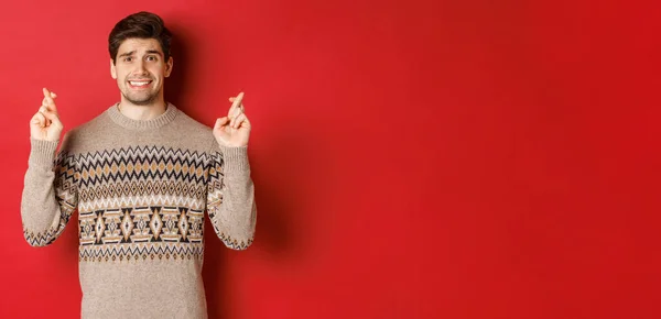 Imagen del hombre esperanzado y preocupado en jersey de Navidad esperando algo, dedos cruzados para la buena suerte y pidiendo deseos, nervioso por el regalo de año nuevo, fondo rojo —  Fotos de Stock