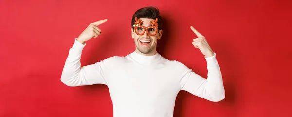 Όμορφος χαρούμενος τύπος με λευκό πουλόβερ, που δείχνει τα χριστουγεννιάτικα γυαλιά του, γιορτάζει το νέο έτος και διασκεδάζει, στέκεται πάνω από το κόκκινο φόντο — Φωτογραφία Αρχείου