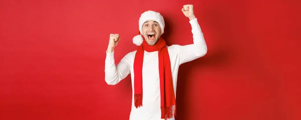 Kép vidám kaukázusi férfi télapó kalap és sál, kiabálva öröm és felemeli a kezét, ünnepli győzelem vagy győzelem, diadalmaskodik a piros háttér — Stock Fotó