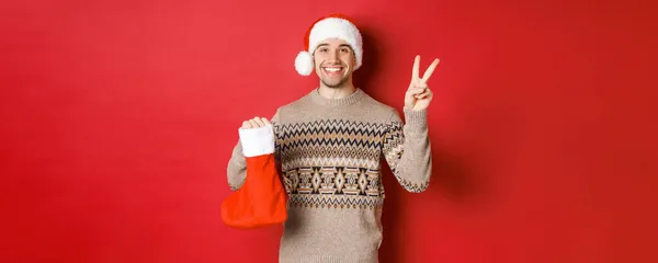 Koncepcja ferii zimowych, Nowy Rok i świętowanie. Obraz szczęśliwego uśmiechniętego mężczyzny w kapeluszu i swetrze Świętego Mikołaja, pokazujący znak pokoju i świąteczną torbę z prezentami, czerwone tło — Zdjęcie stockowe