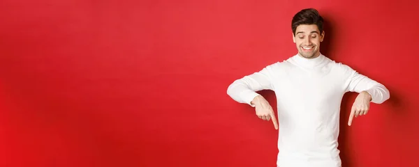 Bild av drömmande snygg man i vit tröja, ser och pekar finger ner på kopieringsutrymme, annons om nyår och helgdagar, står över röd bakgrund — Stockfoto