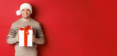 Noel Baba şapkalı ve kışlık süveterli yakışıklı bir adam resmi, bir hediye tutuyor, Noel hediyesi veriyor ve kırmızı arka planda durup mutlu tatiller diliyor.