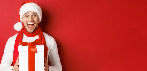 Концепция зимних праздников, Рождества и образа жизни. Крупный план взволнованного красавца в бандаже и шарфе Санты, выглядящего изумленным и получающего новогодний подарок, стоящего на красном фоне — стоковое фото