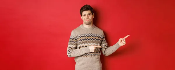 Concept of christmas celebration, winter holidays and lifestyle. Image of skeptical and displeased handsome man in xmas sweater, pointing fingers right and do not recommend product — Stock Photo, Image