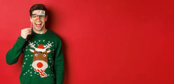 Портрет веселого красивого чоловіка в різдвяному светрі, що тримає смішну вечірню маску і посміхається, святкуючи зимові канікули, стоїть на червоному тлі — стокове фото