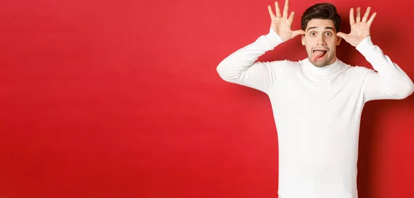 Портрет смешного белого парня, показывающего язык и делающего лица, носящего белый свитер, стоящего на красном фоне — стоковое фото