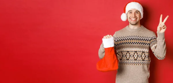 Konzept der Winterferien, Neujahr und Feier. Bild eines glücklich lächelnden Mannes mit Weihnachtsmütze und Pullover, der ein Friedenszeichen und eine Weihnachtsstrumpftasche mit Geschenken zeigt, roter Hintergrund — Stockfoto