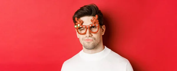 Conceito de férias de inverno, Natal e celebração. Close-up de homem atraente atrevido em óculos de festa, piscando e olhando atrevido, de pé sobre fundo vermelho — Fotografia de Stock