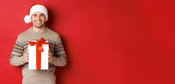 Image d'un bel homme souriant en chapeau de Père Noël et pull d'hiver, tenant un cadeau, donnant cadeau de Noël et souhaitant de bonnes vacances, debout sur fond rouge — Photo