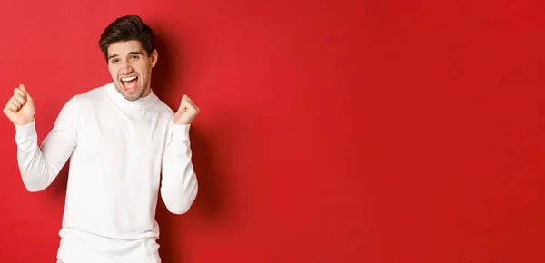Portrait of handsome man enjoying new year party, dancing and having fun, standing in white sweater against red background — Stock Photo, Image