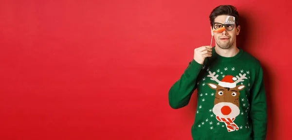 Noel süveteri ve parti maskesi takmış komik bir adam, kış tatilini kutluyor, komik yüzler gösteriyor, kırmızı arka planda duruyor. — Stok fotoğraf