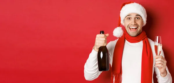 Pojęcie ferii zimowych, Bożego Narodzenia i stylu życia. Zbliżenie wesołego przystojniaka, trzymającego butelkę szampana i szklankę, świętującego Nowy Rok, stojącego na czerwonym tle — Zdjęcie stockowe