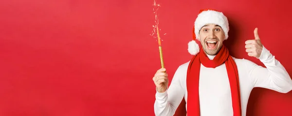 Concetto di Natale, vacanze invernali e celebrazione. Bell'uomo che festeggia il nuovo anno e si diverte, tiene scintillante e mostra pollice in su, in piedi sopra lo sfondo rosso — Foto Stock