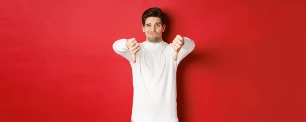 Porträtt av besviken skeptisk kille, klädd i vit tröja, visar tummen ner till oense, uttrycka ogillar, står över röd bakgrund — Stockfoto
