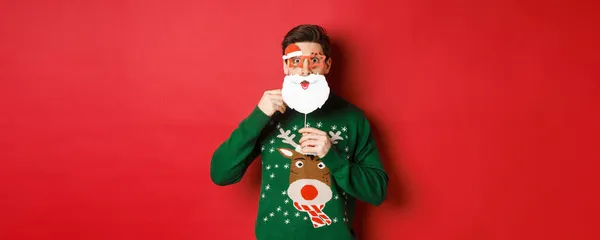 Retrato del hombre sorprendido en jersey de navidad verde, sosteniendo divertida máscara de Santa Claus, celebrando año nuevo, de pie sobre fondo rojo —  Fotos de Stock