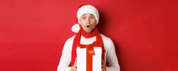 Концепция зимних праздников, Рождества и образа жизни. Крупный план удивленного красавца в шляпе Санты и шарфе, выглядящего изумленным и держащего новогодний подарок, стоящего на красном фоне — стоковое фото