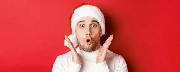 Primo piano di stupito bell'uomo in cappello di Babbo Natale e maglione bianco, alzando le mani e guardando impressionato, in piedi su sfondo rosso — Foto Stock