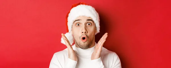 Close-up de homem bonito espantado em chapéu de santa e suéter branco, levantando as mãos e olhando impressionado, de pé sobre fundo vermelho — Fotografia de Stock