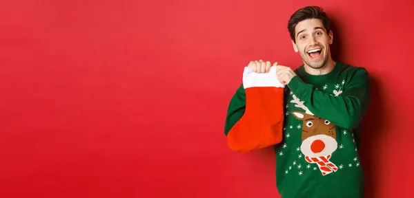 Зображення привабливого веселого чоловіка в светрі, що тримає різдвяні панчохи з подарунками, святкуючи зимові канікули, стоячи на червоному тлі — стокове фото