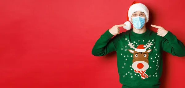 Az új év koncepciója, a koronavírus és a társadalmi távolság. Boldog ember télapó kalap és karácsonyi pulóver, azt javasolja, hogy viseljen orvosi maszkot a party, álló felett piros háttér — Stock Fotó