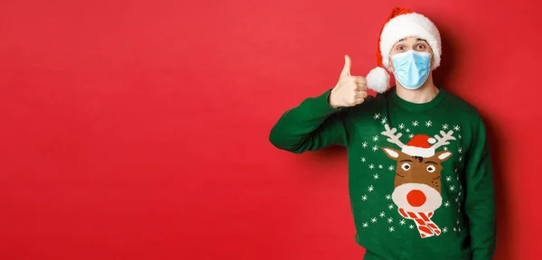 Yeni yıl kavramı, covid-19 ve sosyal mesafe. Şaşırmış genç adam Noel 'i kutluyor, tıbbi maske takıyor ve Noel Baba şapkası takıyor, kırmızı arka planda baş parmağını kaldırıyor. — Stok fotoğraf