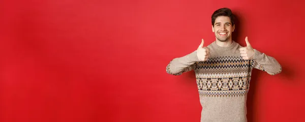 Ritratto di ragazzo bello felice e contento in maglione di Natale, mostrando pollici in su e annuire in approvazione, sorridente soddisfatto, in piedi su sfondo rosso — Foto Stock