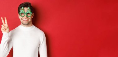 Beyaz süveter ve gözlüklü yakışıklı bir adam resmi. Barış işareti ve gülümseme gösteriyor. Mutlu Noeller diliyor.