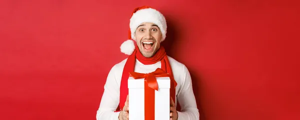 Concept of winter holidays, christmas and lifestyle. Close-up of excited handsome man in santa jat and scarf, looking amazed and receiving new year present, standing over red background — Stock Photo, Image