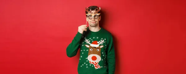 Αστείος άνθρωπος με χριστουγεννιάτικο πουλόβερ και μάσκα κόμμα, γιορτάζει τις χειμερινές διακοπές, δείχνει αστεία πρόσωπα, στέκεται πάνω από το κόκκινο φόντο — Φωτογραφία Αρχείου
