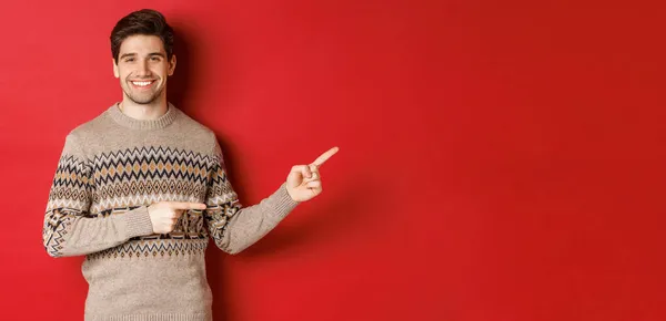 Obrázek atraktivního usmívajícího se muže, na sobě vánoční svetr, ukazující prsty vpravo a ukazující novoroční reklamu, stojící nad červeným pozadím — Stock fotografie
