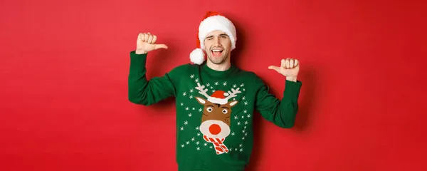 Koncept zimních prázdnin, vánoc a životního stylu. Drzý pohledný muž v Santa klobouku a zeleném svetru, ukazuje na sebe a mrká, stojí nad červeným pozadím — Stock fotografie