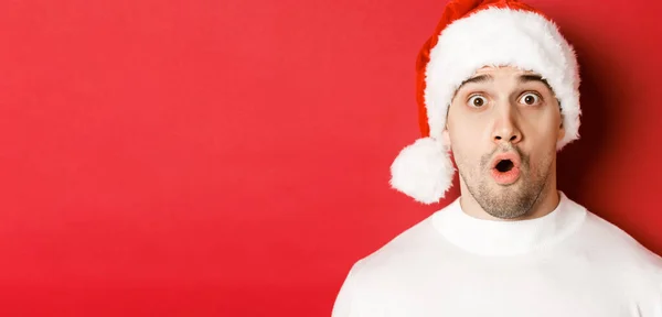 Nahaufnahme eines attraktiven, überraschten Mannes mit Weihnachtsmütze, der wow sagt und etwas Interessantes betrachtet, vor rotem Hintergrund steht — Stockfoto