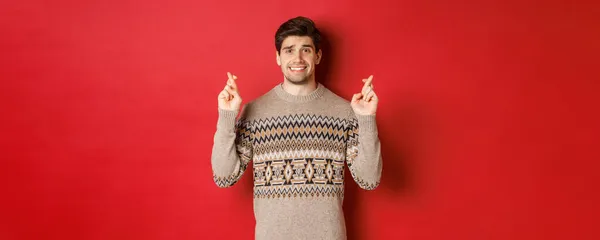 Immagine di uomo speranzoso e preoccupato in maglione di Natale in attesa di qualcosa, incrociare le dita per buona fortuna e fare il desiderio, nervoso per il regalo di Capodanno, sfondo rosso — Foto Stock