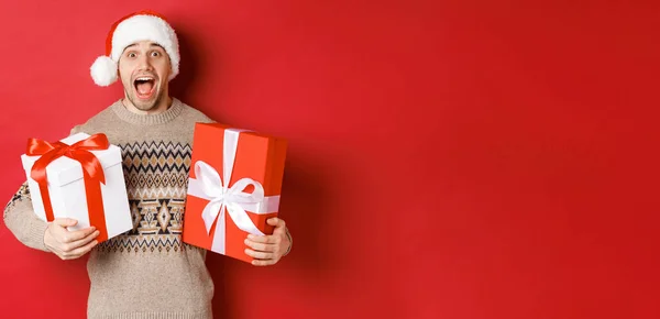 Bild eines fröhlichen attraktiven Kerls mit Weihnachtsgeschenken, der mit Weihnachtsmütze und Winterpullover vor rotem Hintergrund steht und erstaunt lächelt — Stockfoto